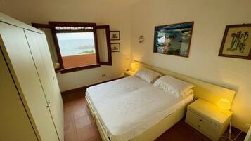 A bedroom with a bed, a nightstand, a window with a view of the sea, and a wardrobe.