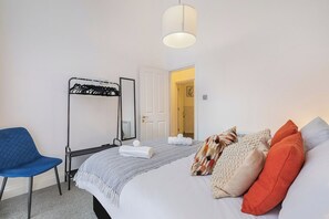 Habitación de hotel con una cama, una silla, perchero y una puerta que da a otra habitación.