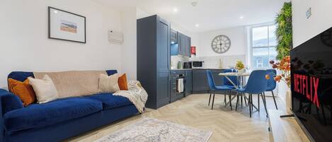 Un soggiorno moderno con un divano blu, un'area pranzo, una cucina e uno schermo TV che mostra 'NETFLIX'.