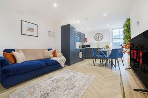 A modern living space with a blue sofa, a dining area, a kitchen, and a TV screen displaying 'NETFLIX'. - Central Brighton 2BR | Sleeps 4 | Near Seafront (Brighton and Hove)