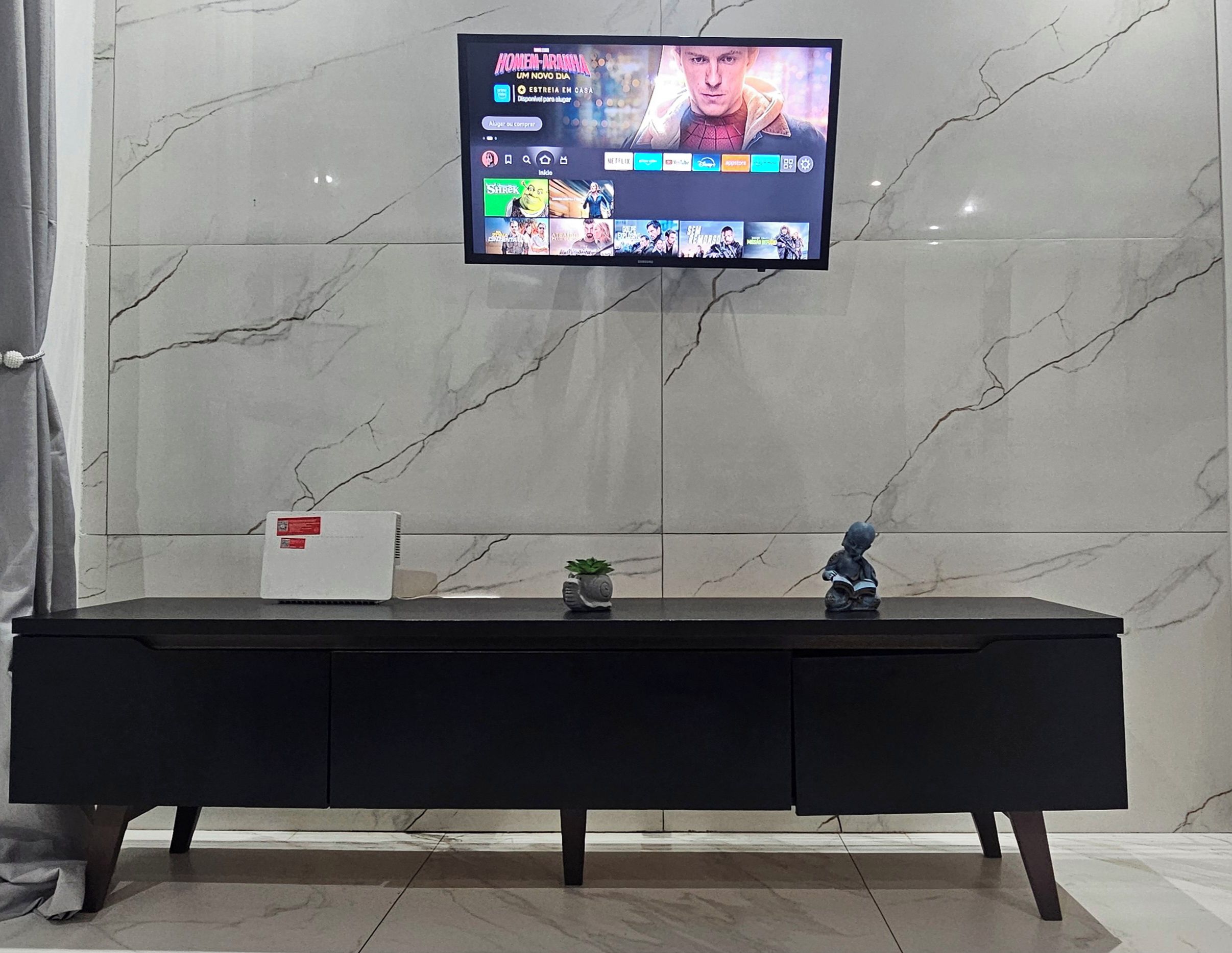 A television mounted on a wall with a marble pattern, a black desk with drawers, and a small plant on the desk.
