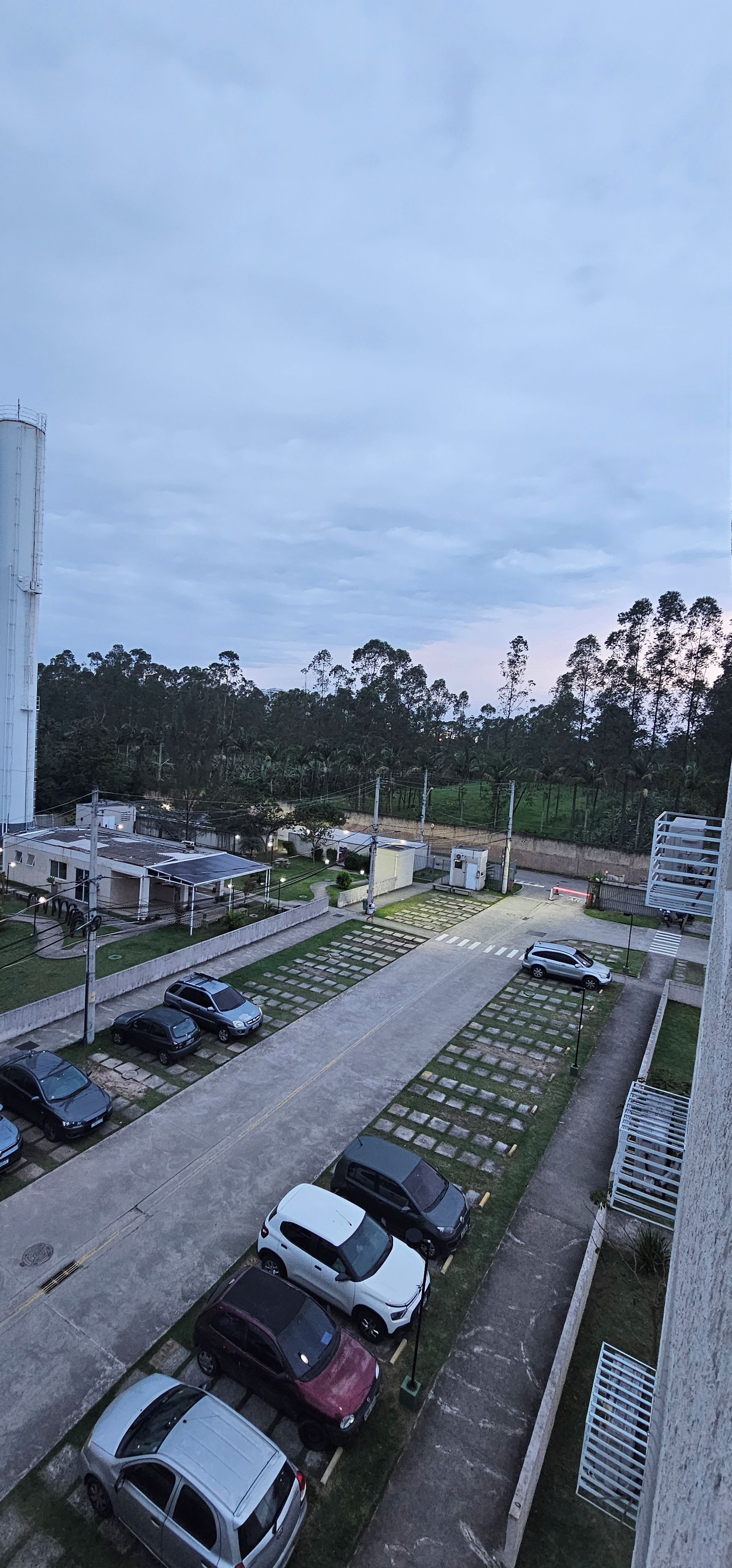 A parking lot with several cars, a building with a flat roof, and a grassy area with trees in the background.