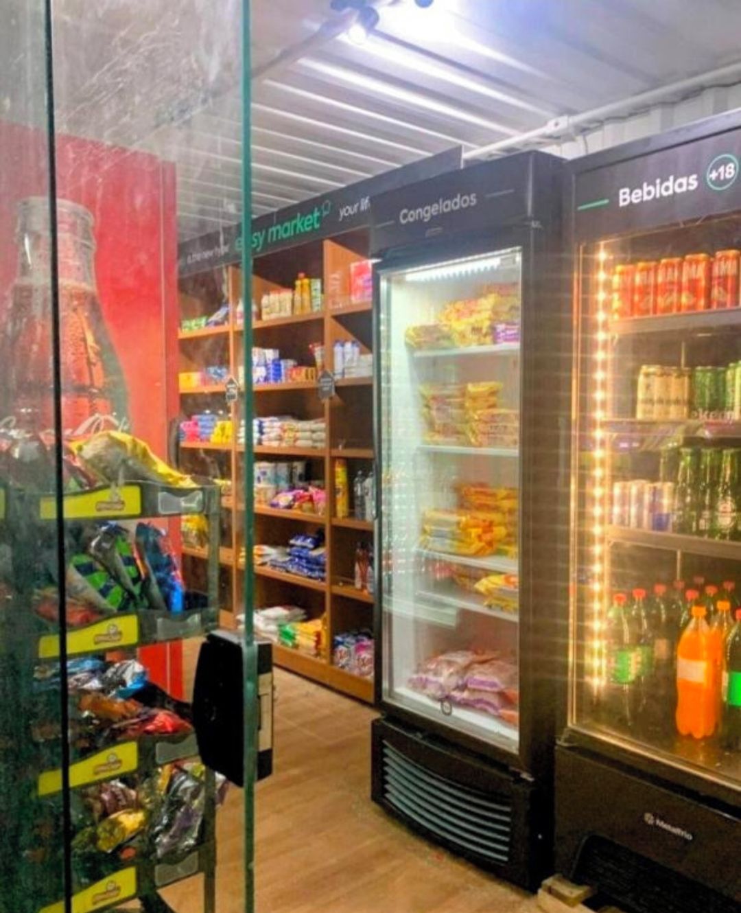 A refrigerated display area with snacks and beverages, shelves with packaged goods, and a glass door entrance.