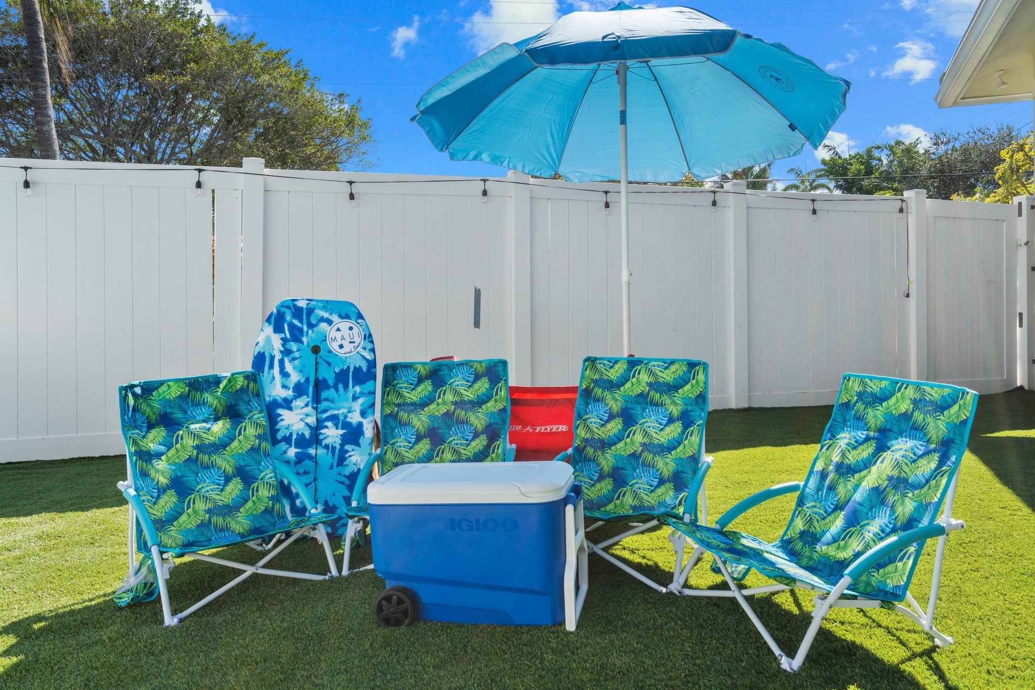 Un jardin avec une clôture blanche, un parasol bleu et quatre chaises de plage ornées d’un motif de feuilles tropicales.