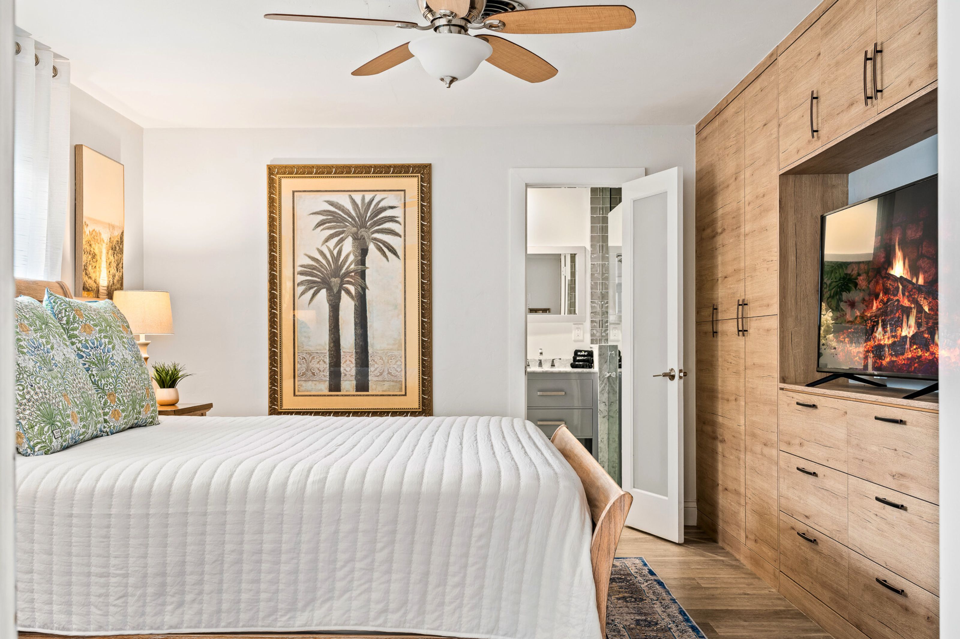 Une chambre à coucher comprenant un lit, un ventilateur de plafond, un tableau encadré, un téléviseur et une armoire en bois.
