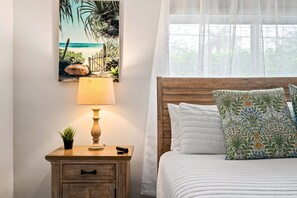 A bedroom with a wooden headboard, a bed with white bedding, a nightstand with a lamp, a small plant, and a framed picture of a beach scene. - Walk to Beach & Restaurants | Hot Tub | Beach Gear (Pompano Beach)