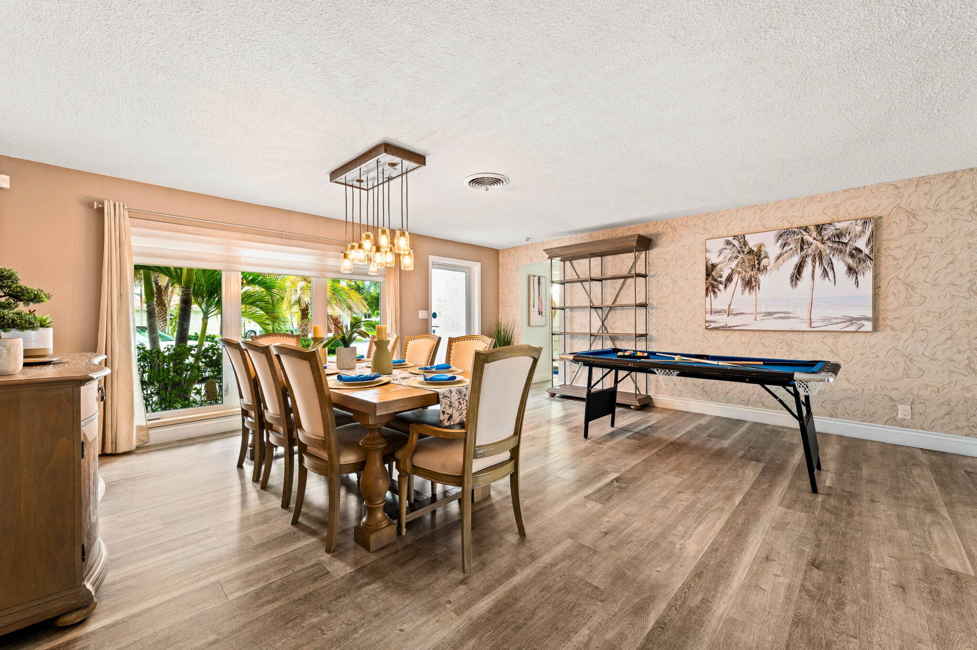 A dining area with a pool table, wooden flooring, and a view of greenery through large windows.
