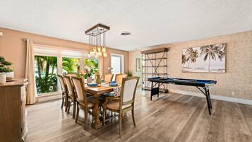 A dining area with a pool table, wooden flooring, and a view of greenery through large windows.