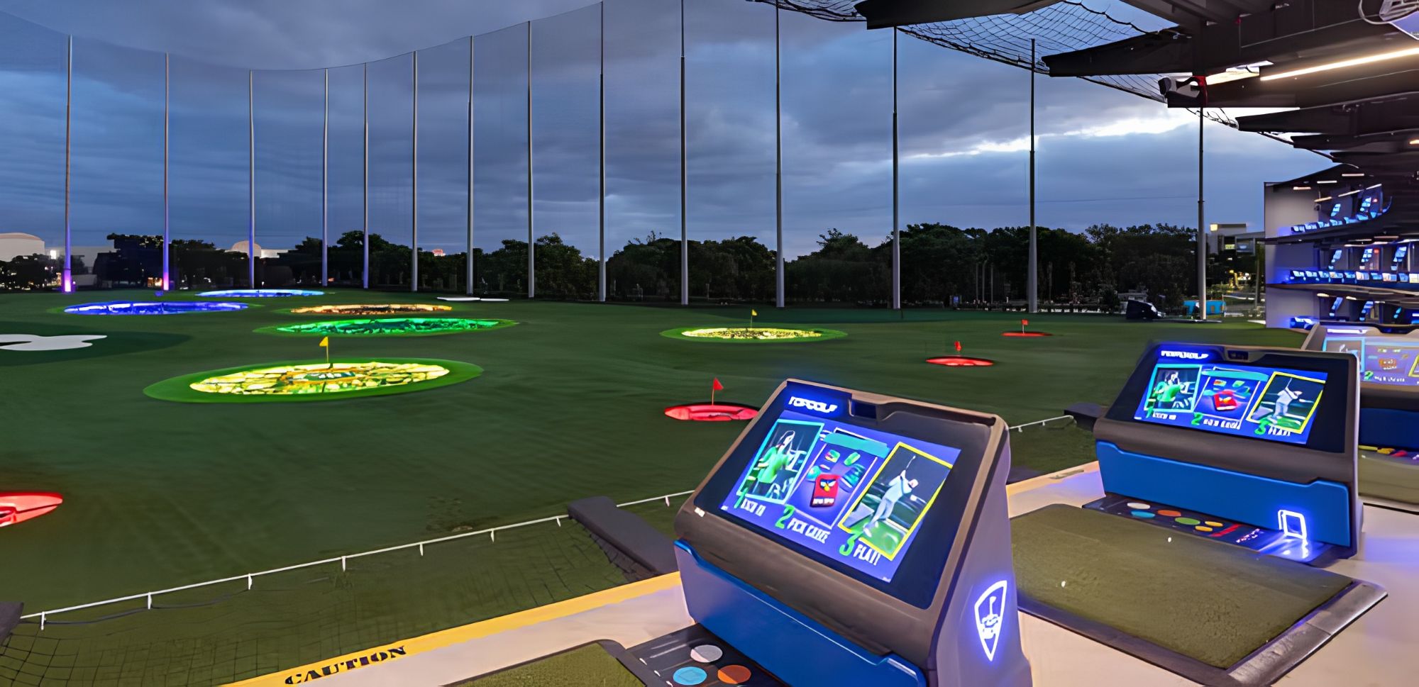 A golf simulation setup with multiple screens displaying game interfaces, a green artificial turf, and a netted overhead structure.