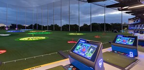 Ein Golf-Simulationssystem mit mehreren Bildschirmen, die Spielschnittstellen zeigen, einem grünen Kunikrasen und einer überdachten Netzkonstruktion.