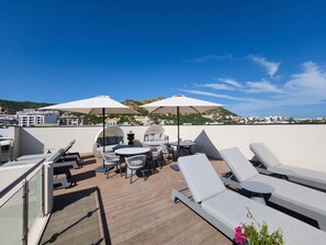 Terraço no telhado com espreguiçadeiras, mesa e guarda-sóis, com vista para a paisagem urbana.