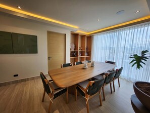 Une salle à manger moderne avec une table et des chaises en bois, une bibliothèque intégrée et une grande fenêtre ornée de rideaux légers.