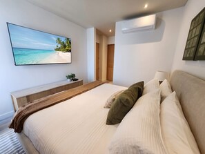 Ein Schlafzimmer mit einem Bett, Kissen, einem Nachttisch, einem an der Wand befestigten Fernseher und einer Klimaanlage.
