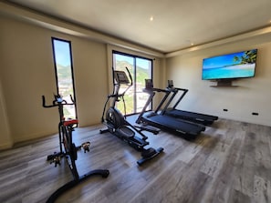 Ein Heimfitnessraum mit Trainingsgeräten, großen Fenstern und einem Fernseher.