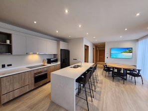 A modern kitchen with a central island, a dining area with a large TV, and a wooden floor. - Ameth: Spacious 3BR Apartment with Rooftop Pool (Cabo San Lucas)