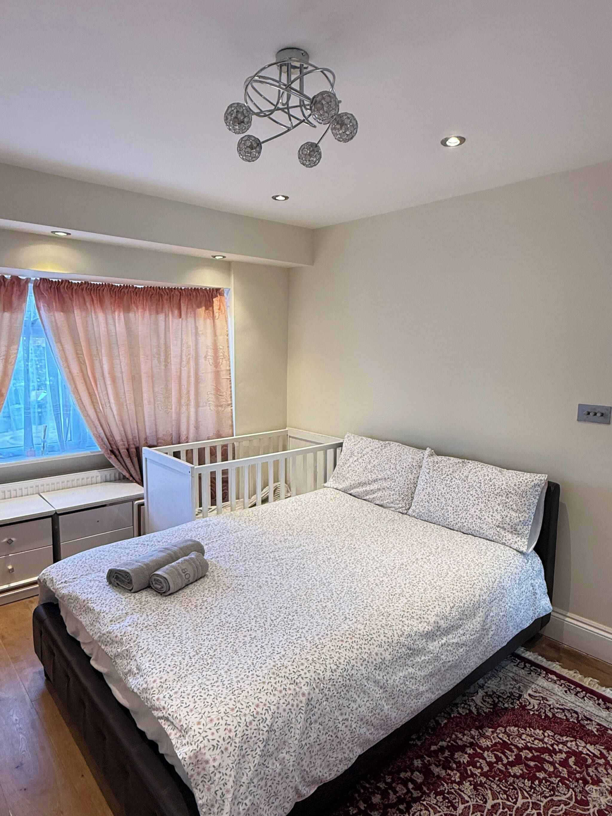 Une chambre avec un lit simple, un berceau, une fenêtre avec des rideaux et un luminaire au plafond.