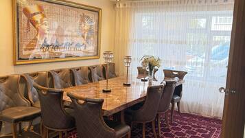 Un espace repas avec une longue table, des chaises rembourrées, un lustre et une grande œuvre d’art encadrée au mur.