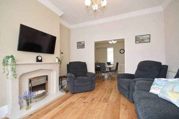 A living room with a fireplace, two grey armchairs, a sofa, a flat-screen TV, and a dining area in the background. - Sea Escape - Convenient location (North Yorkshire)