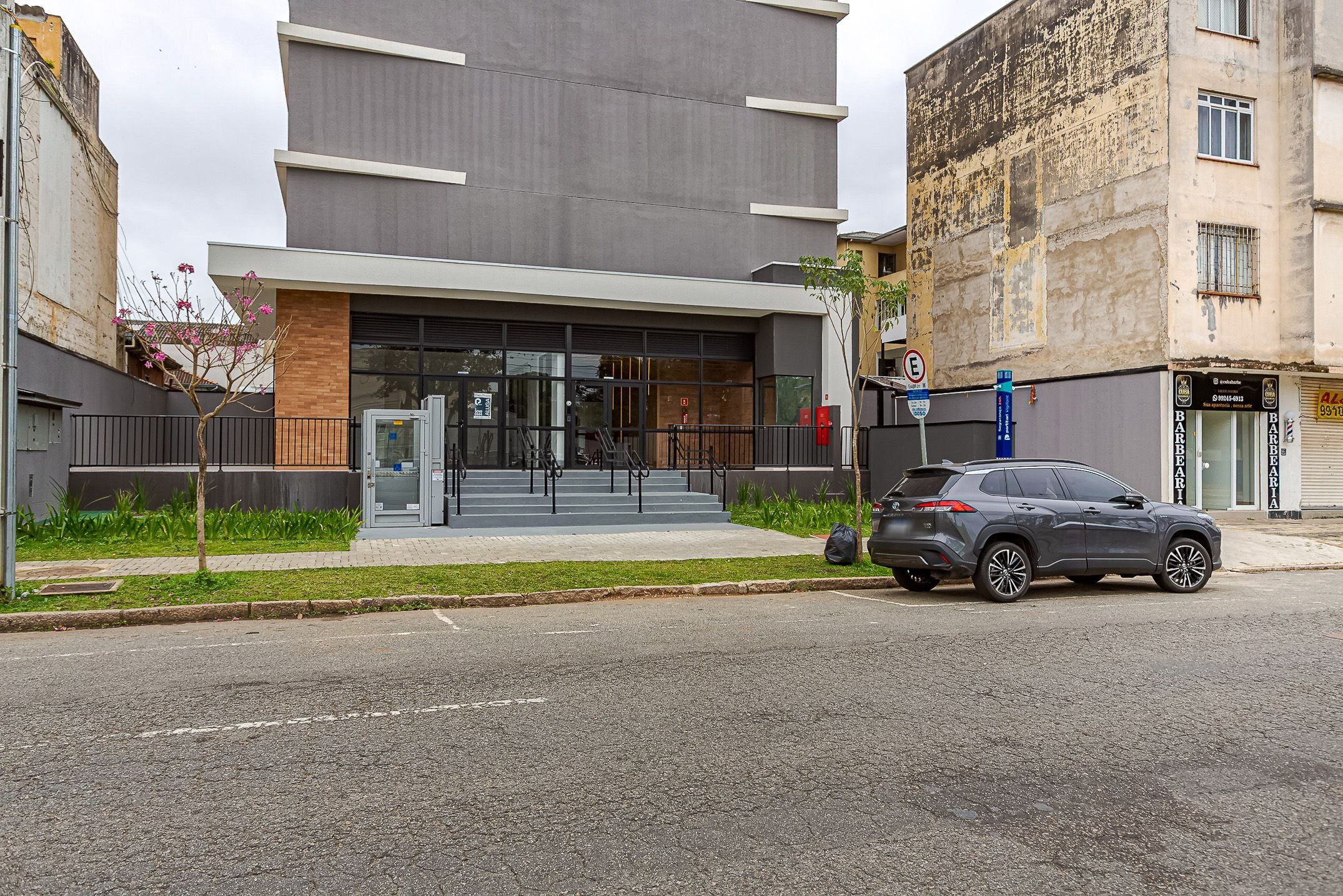 A modern building with a glass entrance, a parking area, and a street with a parked car.