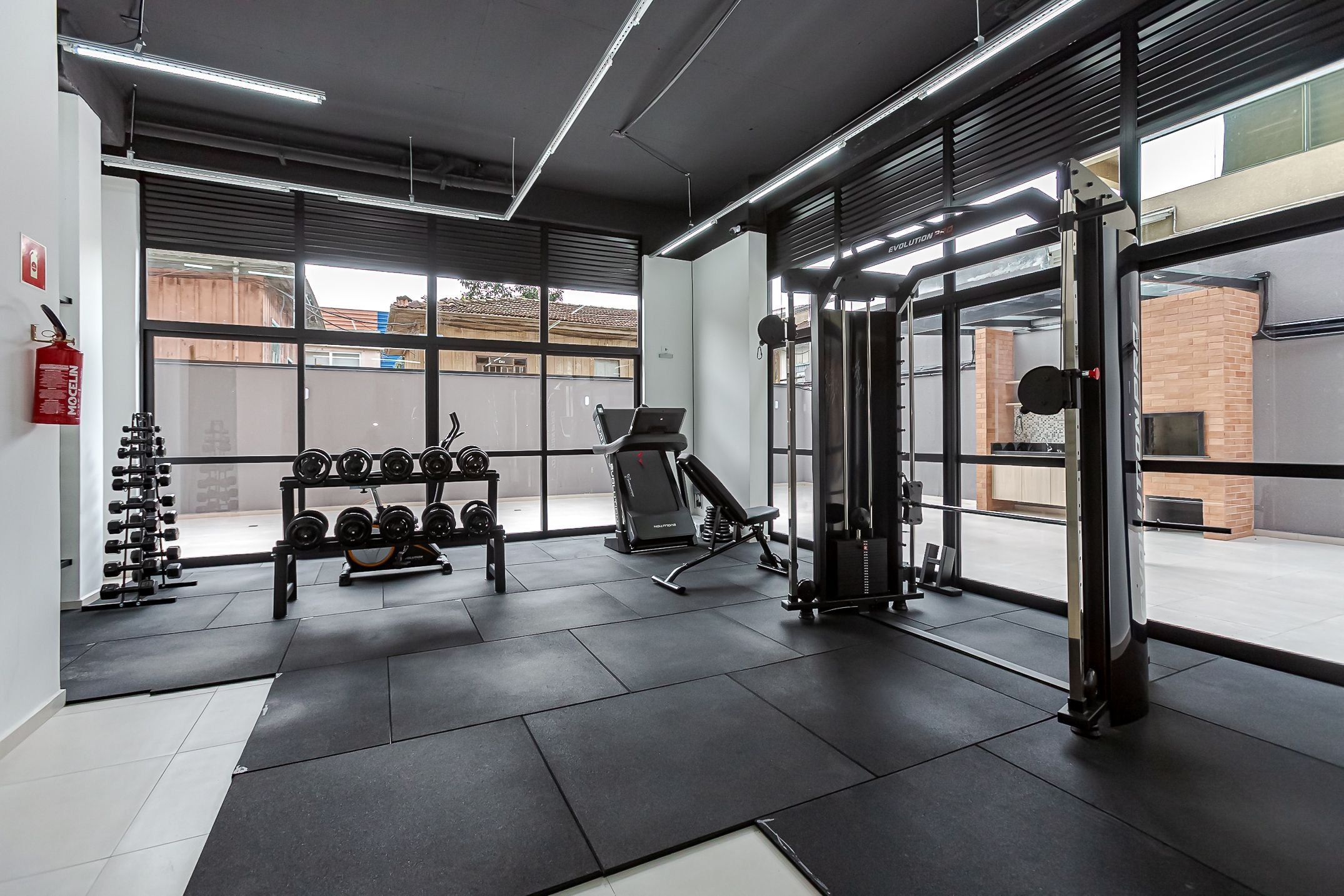 Un gimnasio con equipamiento de ejercicio, un estante para mancuernas y ventanas amplias.