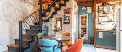 Un interior rústico con una escalera de madera, un comedor con sillas, una cocina con un fregadero y una pared de piedra.
