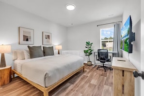 Une chambre moderne avec un grand lit, des lampes de chevet, un bureau avec une chaise, une plante en pot et un téléviseur fixé au mur.