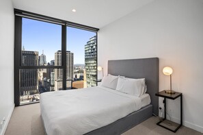 Ein Hotelzimmer mit einem großen Bett, einem Nachttisch, einer Lampe und Blick auf die Stadt durch große Fenster.