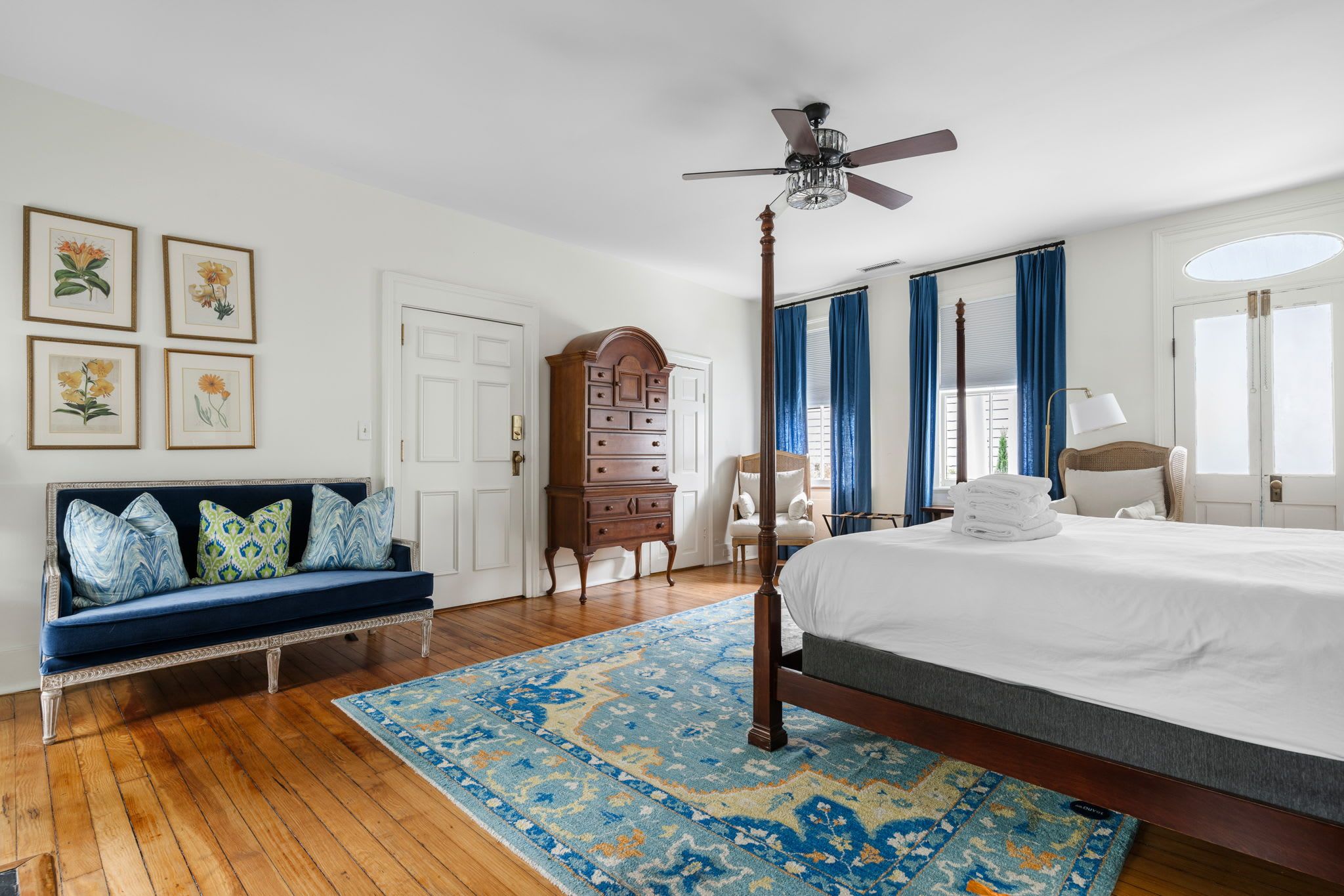 A bedroom with a large bed, a sitting area with a bench, a wardrobe, and a ceiling fan.