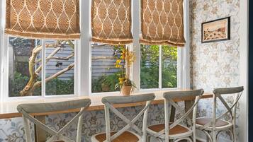 Une pièce comprenant un coin repas avec une table en bois et quatre chaises, de grandes fenêtres ornées de rideaux à motifs et un plafond agrémenté d’éléments décoratifs.