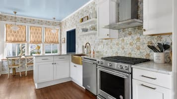 Une cuisine avec des meubles blancs, une cuisinière en acier inoxydable et une fenêtre ornée de rideaux à motifs.