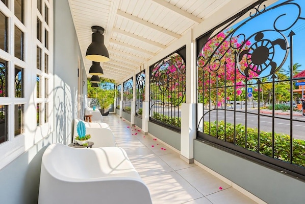 A covered outdoor seating area with white chairs, a black pendant light, and a view of a street with pink flowers.