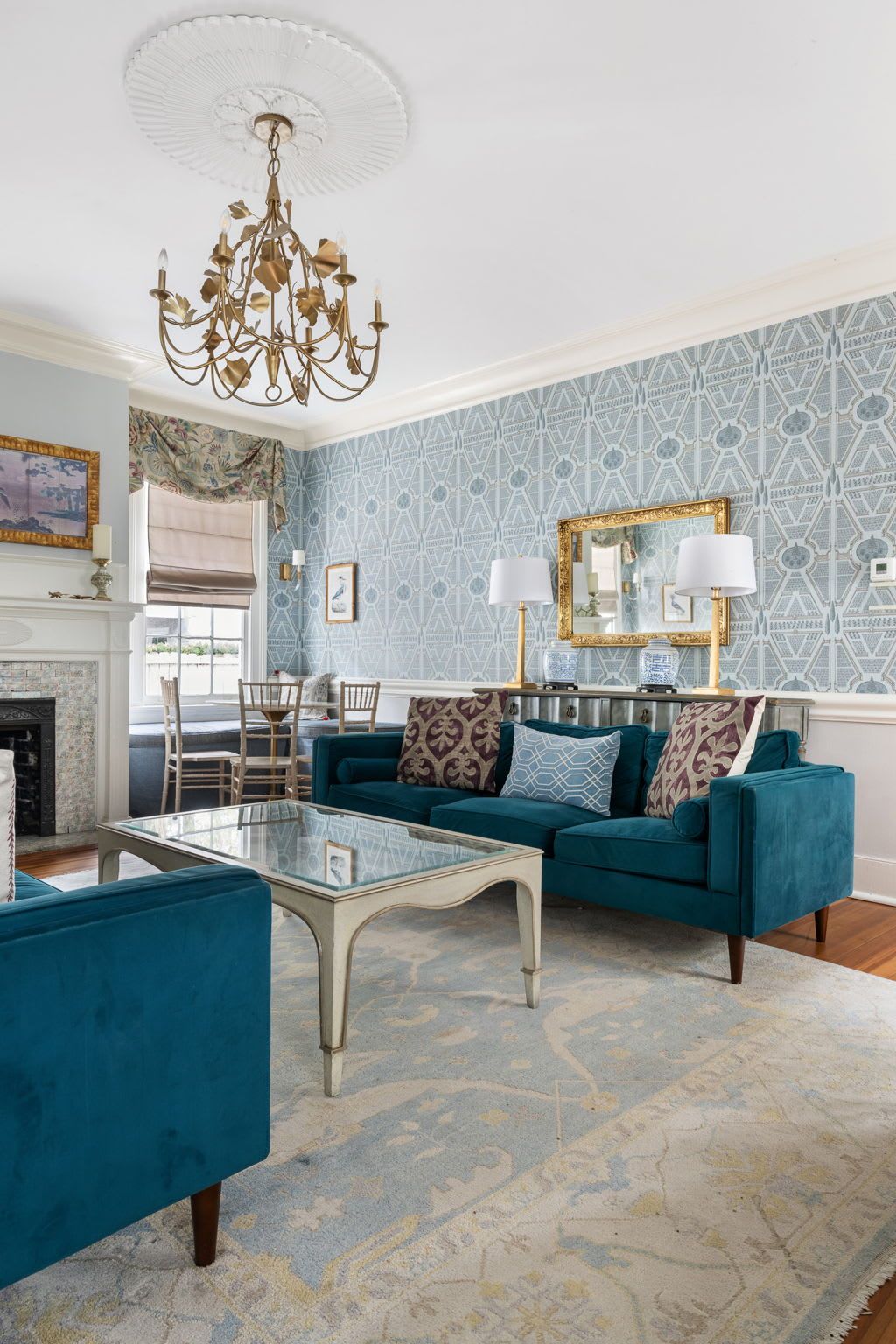 A living room with a blue sofa set, a glass coffee table, a chandelier, and patterned wallpaper.