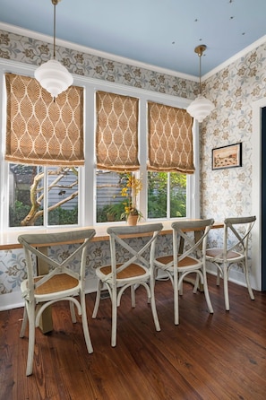 Quarto com área de refeições, mesa de madeira e quatro cadeiras, janelas amplas com cortinas estampadas e teto com detalhes decorativos.