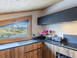 A modern kitchen with wooden cabinets, a black countertop, a window with a view of a mountainous landscape, and a vase with flowers on the counter. - Cozy apartment near the slopes - 4 guests (Belle Plagne)