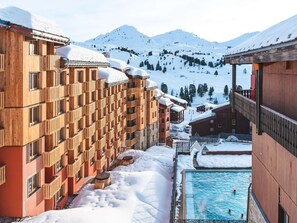 A snow-covered mountain resort with wooden buildings, a pool area, and a clear view of the mountains. - Cozy apartment near the slopes - 4 guests (Belle Plagne)