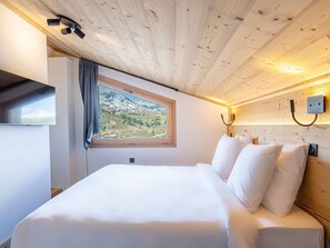 A bedroom with a wooden ceiling, a large window with a mountain view, a bed with white bedding, and a wall-mounted light fixture. - Cozy apartment near the slopes - 4 guests (Belle Plagne)