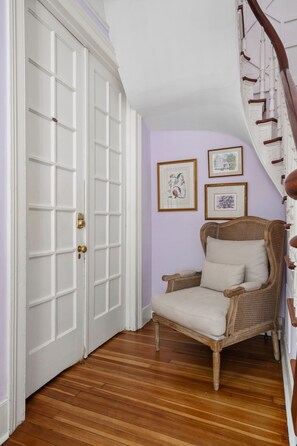 A room with a white door, a wooden floor, a wicker chair, and a staircase. - Bee & Blossom | Large Bed & Breakfast Buyout (Charleston)