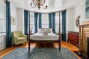 A four-poster bed with a white bedspread, a green armchair, a wooden dresser, and a chandelier. - Bee & Blossom | Large Bed & Breakfast Buyout (Charleston)