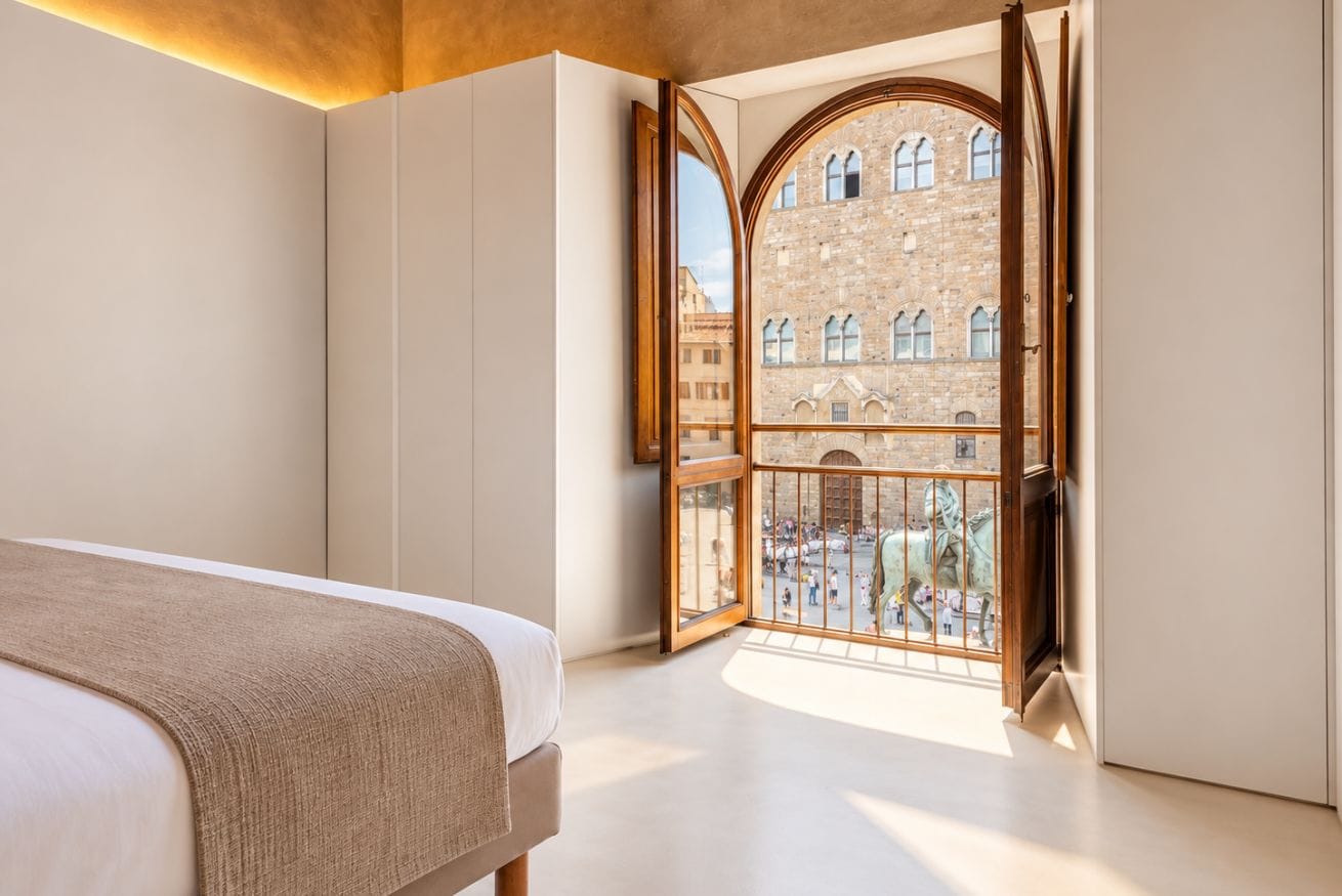 A bedroom with a large bed, a view of a historic building through an arched window, and a wooden door.