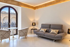 A modern living room with a grey sofa, patterned armchairs, a small round table, and a lamp. A large arched window offers a view of a historic building. - Apartments Florence - Signoria Farine Panoramic view (Firenze)