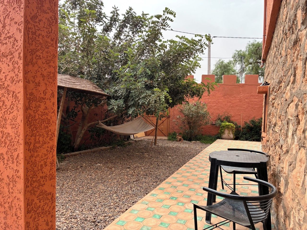 A patio area with a table and chairs, a hammock, and a garden with trees and a brick wall.