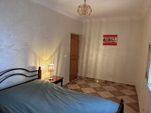 A bedroom with a bed, a nightstand, a lamp, a door, and a checkered floor. - Vrbo Property (Lagfifat)