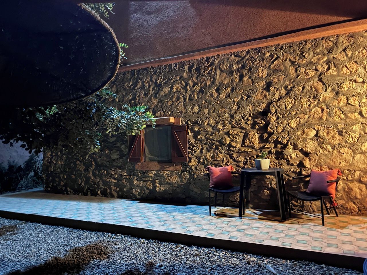 Een terras met een achtergrond van een stenen muur, een tafel met stoelen en een raam met luiken.