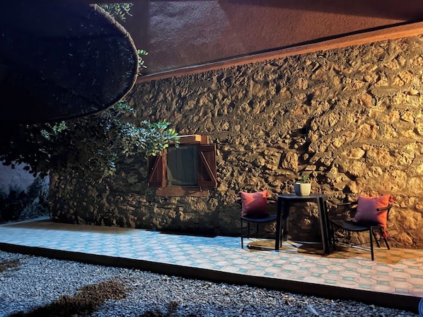 An outdoor seating area with a stone wall backdrop, a table with chairs, and a window with shutters.