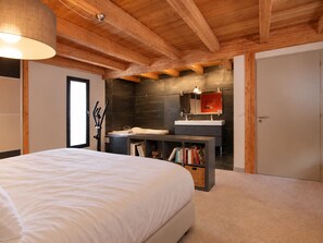 A bedroom with a large bed, a bedside table, a bookshelf, a bathroom with a sink and mirror, and a door. - Contemporary chalet with jacuzzi - 12 guests (Les Deux Alpes)