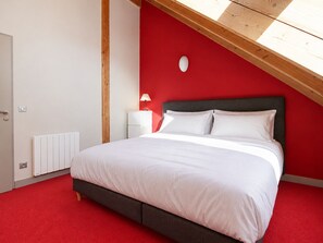 Ein Schlafzimmer mit schräg laufender Decke, einem Bett mit weißer Bettwäsche, einem Nachttisch mit Lampe und einer roten Akzentwand.