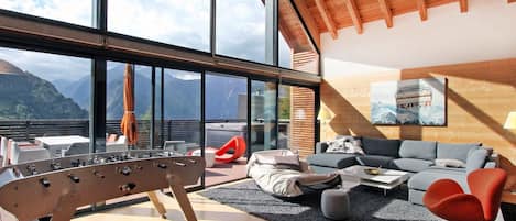 A modern living room with a wooden ceiling, large windows offering mountain views, a sofa set, and a foosball table.