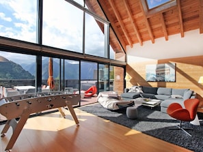 Ein modernes Wohnzimmer mit Holzboden, großen Fenstern mit Blick auf die Berge, einer Sofagarnitur und einem Kicker.