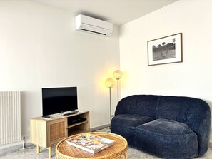 A modern living room with a flat-screen TV, a dark blue sofa, a wicker coffee table with a book, a wall-mounted air conditioner, and a floor lamp. - Air-conditioned apartment, stunning views - 2 guests (Hyères)