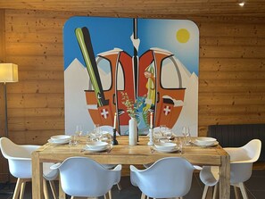 A dining area with a wooden table set for four, white chairs, and a large painting on the wall depicting a mountain scene with a sun. - Semi-detached chalet, mountain view - 6 guests (Méribel)